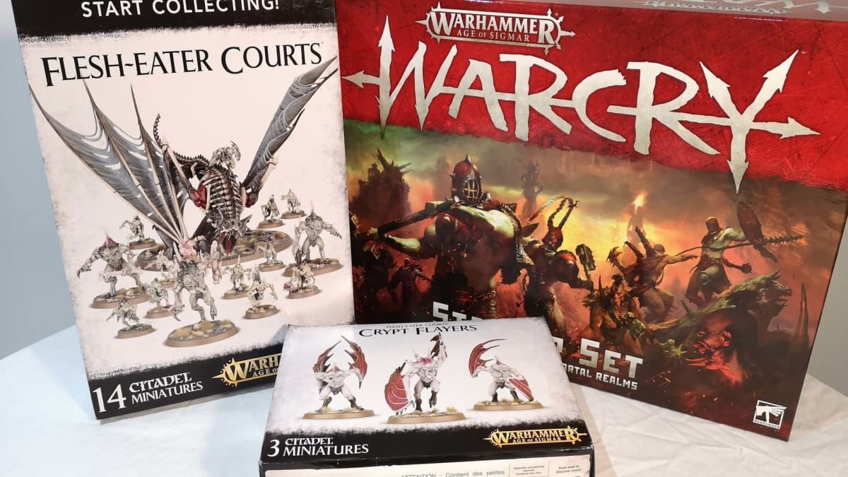 photo showing several boxes for the game WarCry with various miniatures sat on the table in front of the boxes. 