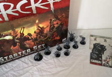 Photo showing several miniatures sitting in front of a warcry box on a table with a white tablecloth placed on top. 