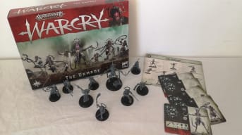 Photo showing a box of WarCry with various miniatures set out on a white table in front of the box. 