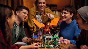 Promotional image of the Lego Ideas Dungeons & Dragons set Red Dragons Tale, showing a group of people at a table. Sitting on the table is the Lego set, showing a tavern, monsters, and a dragon.