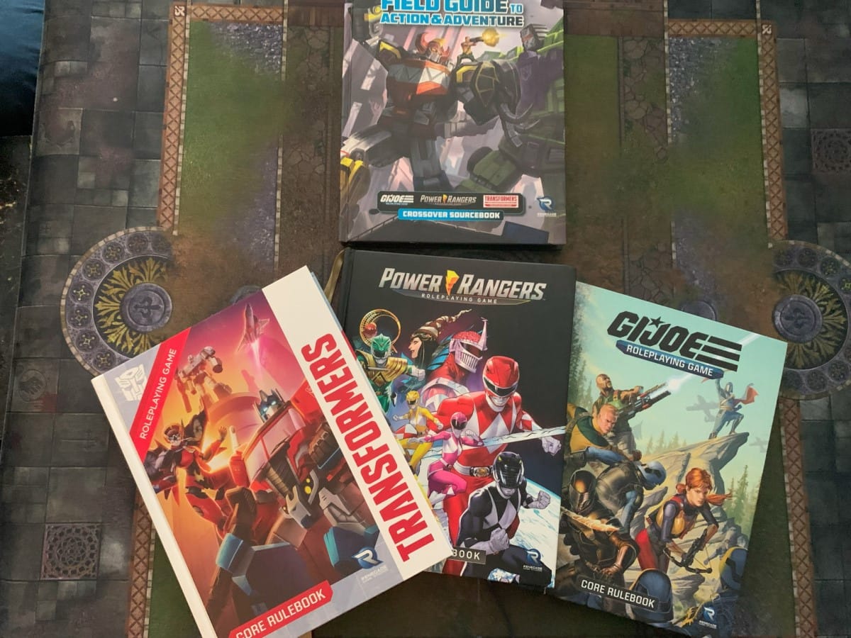 The Essence20 Field Guide to Action & Adventure book on a table spread that includes the core rulebooks for Power Rangers, GI Joe, and Transformers