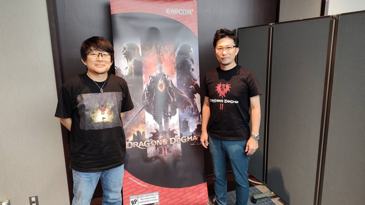 Dragon's Dogma 2 director Hideaki Itsuno and producer Yoshiaki Hirabayashi stand in front of a banner featuring their game's art. 