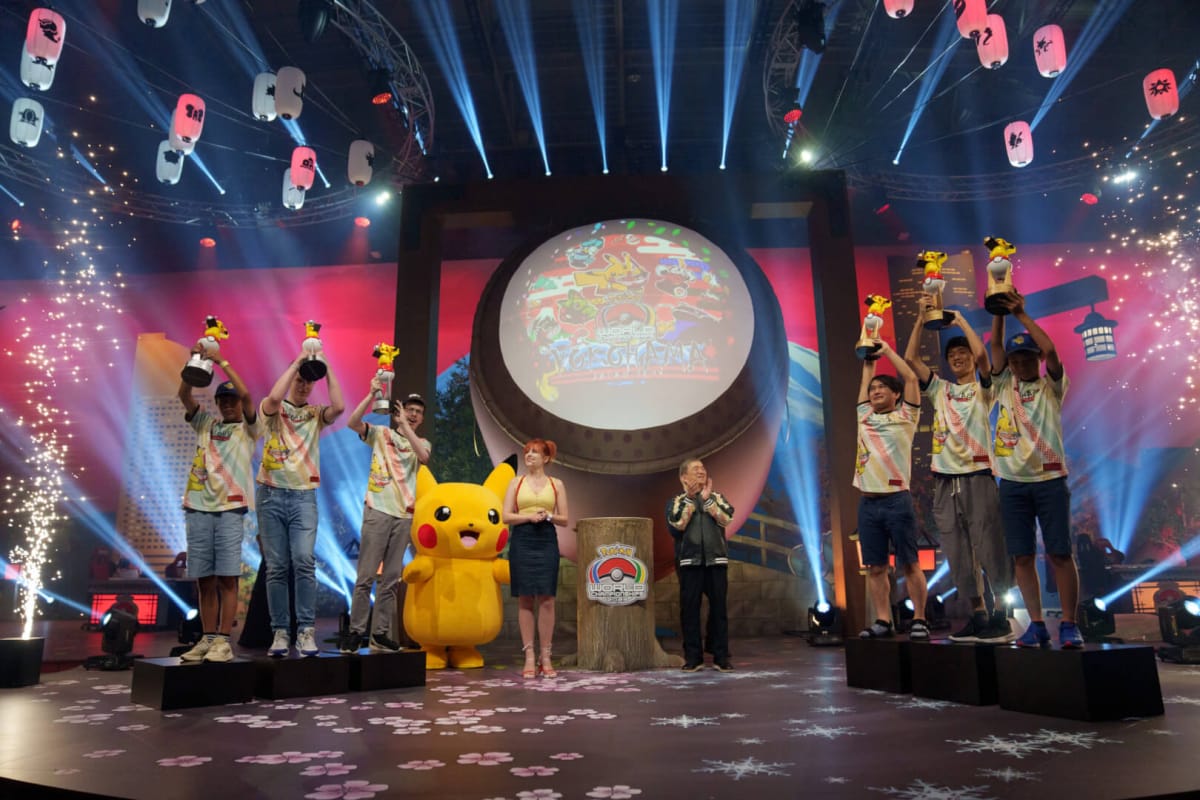 Several players holding aloft Pikachu trophies while hosts look on and smile at the Pokemon World Championships 2023 winners' ceremony