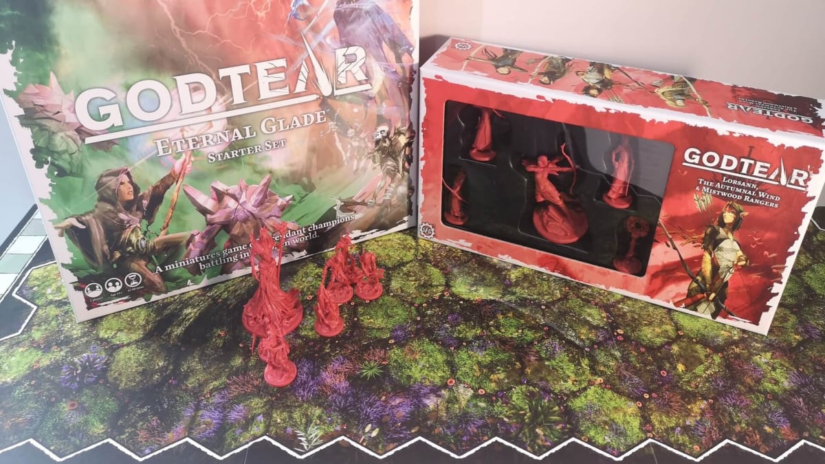 Photo showing several Godtear boxes sitting on a board with several red miniatures sitting on the tabletop nearby. 