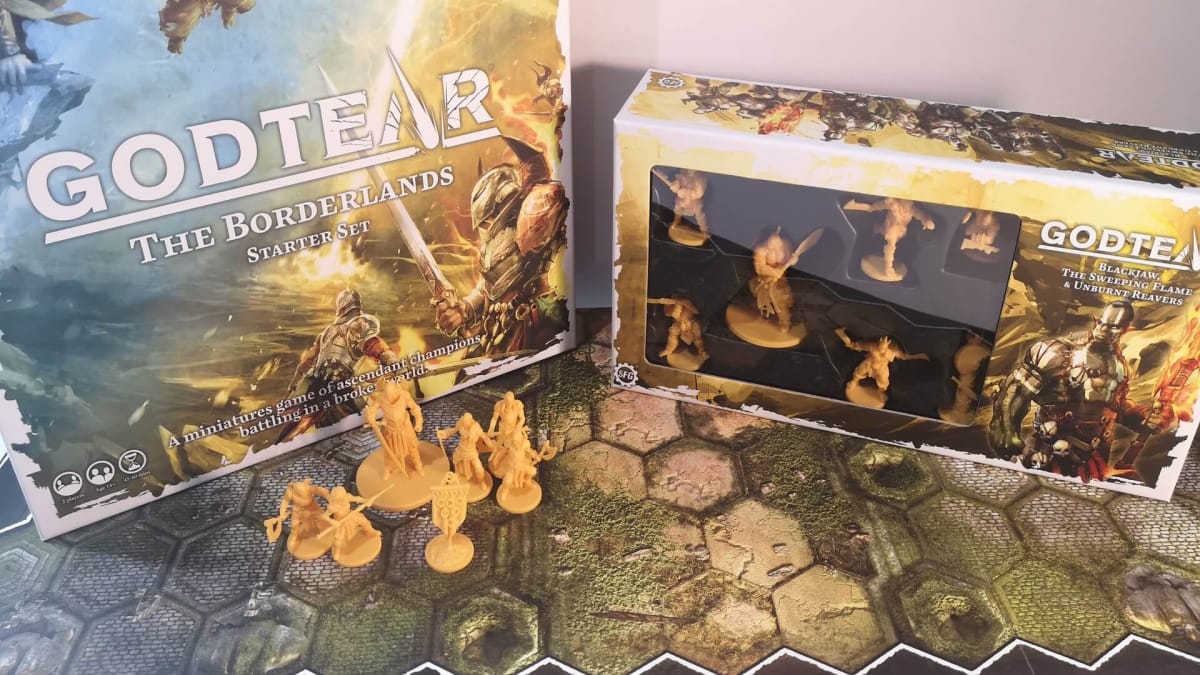 photo showing several Godtear boxes spread out on a table, in front of the boxes a set of miniatures sit on top of the game's board itself. 