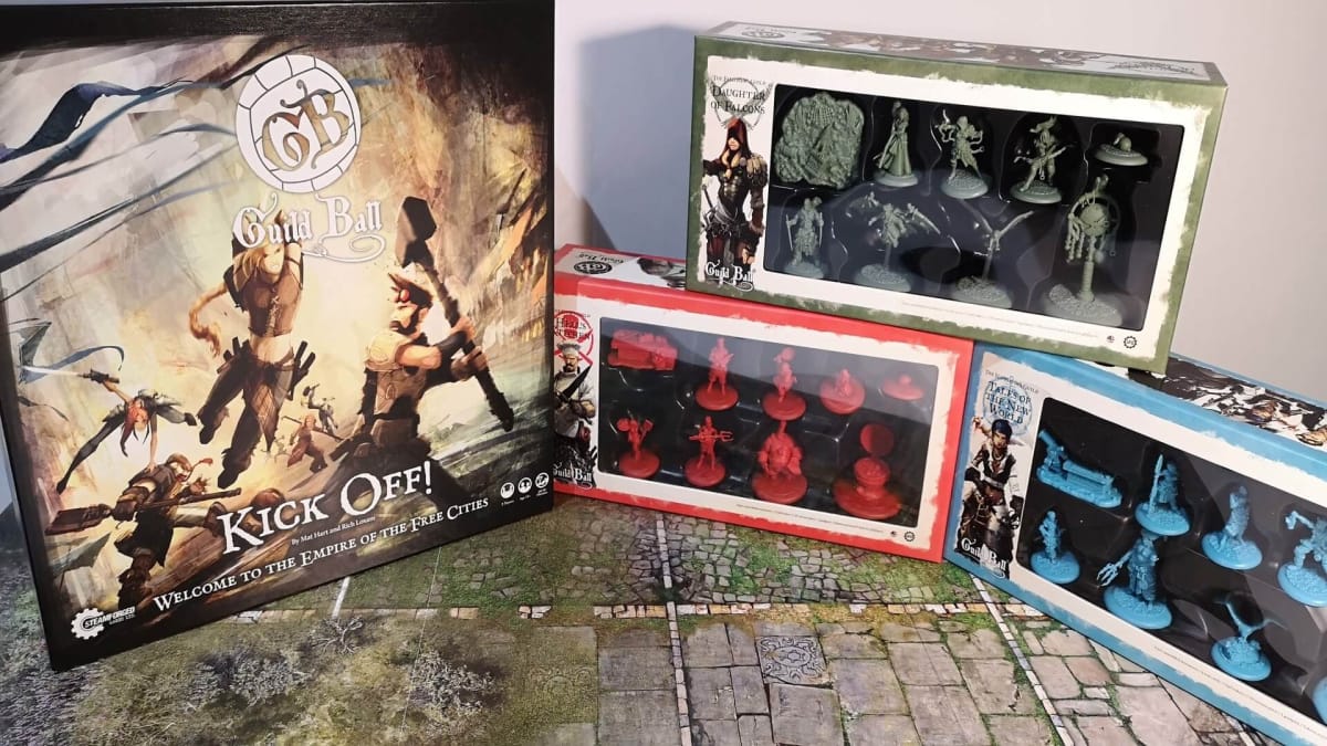Photo showing several boxes of miniatures for the game Guild Ball on a table with the gameboard bellow. 
