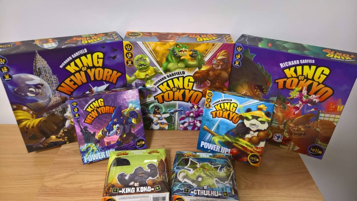Photograph shows several board game boxes of different sizes sitting on a lightwood table with a white wall background. Each of the boxes features cartoonish depictions of various Kaiju-style monsters and city scapes being destroyed around them. 