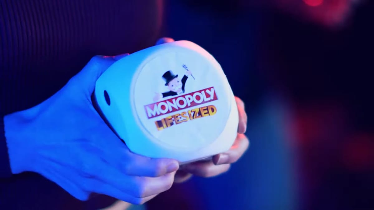 A screenshot of a woman holding a giant die with Monopoly Lifesized written on it