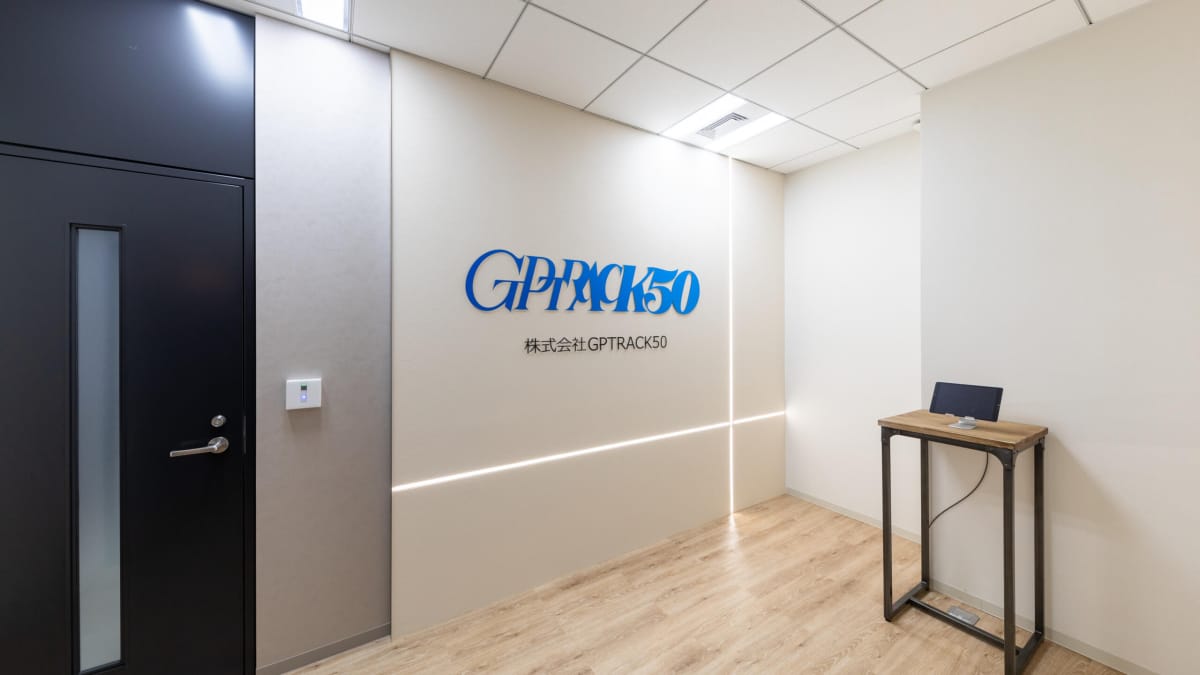 The office of the new Hiroyuki Kobayashi studio GPTRACK50, depicting a fairly sparse room with a company logo, a door, and a device on a table