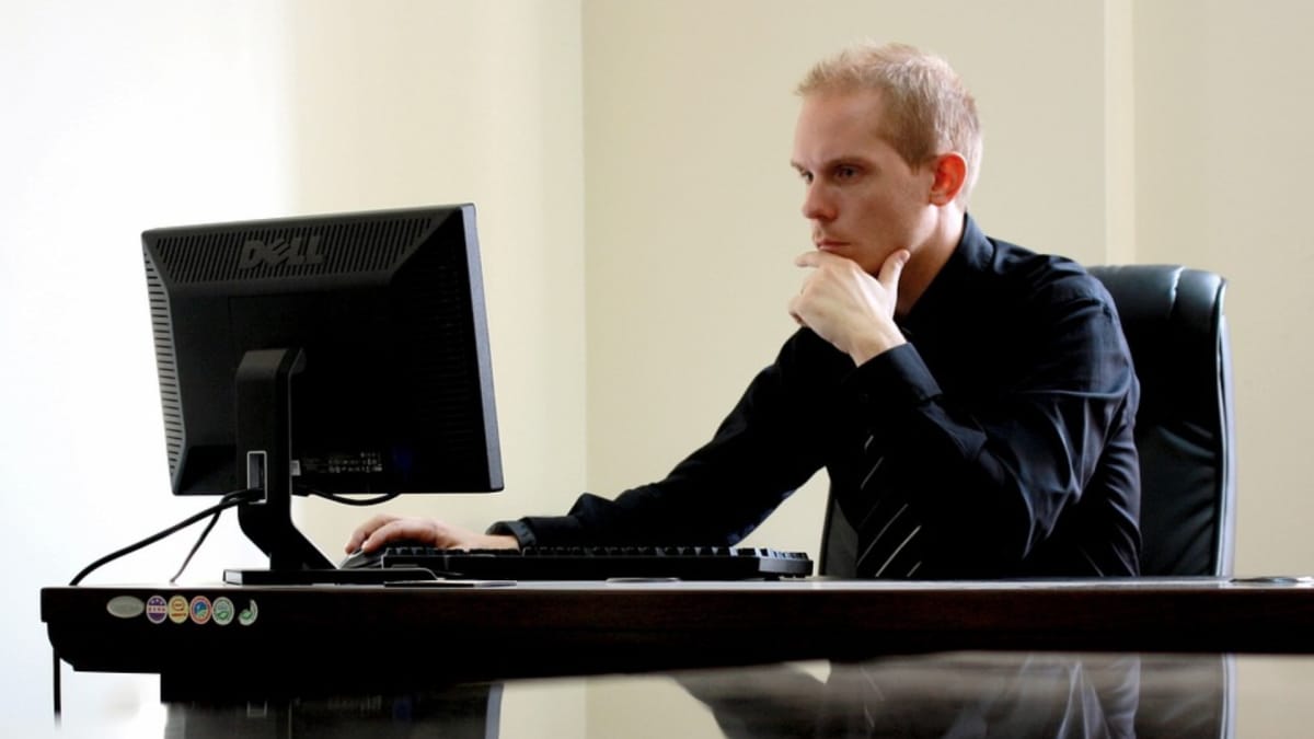 photo showing a man sat looking at a computer screen with a hand thoughtfully on his chin. 