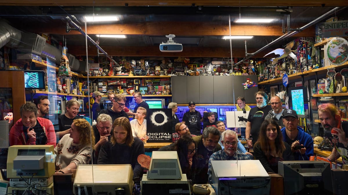 The staff of Digital Eclipse gathered in an office cluttered with gaming merchandise while one of them holds a company logo poster