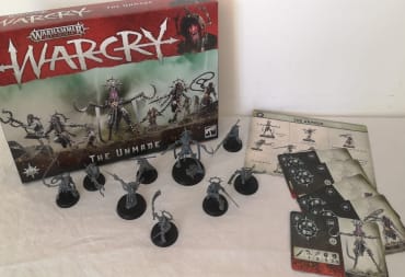 Photo showing a box of WarCry with various miniatures set out on a white table in front of the box. 