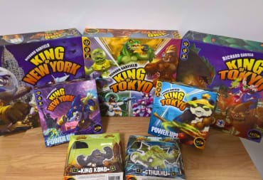 Photograph shows several board game boxes of different sizes sitting on a lightwood table with a white wall background. Each of the boxes features cartoonish depictions of various Kaiju-style monsters and city scapes being destroyed around them. 