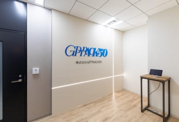 The office of the new Hiroyuki Kobayashi studio GPTRACK50, depicting a fairly sparse room with a company logo, a door, and a device on a table